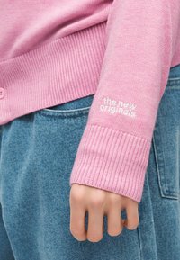 Cilvēka roka blakus gurnam valkā rozā adītu džemperi ar tekstu "the new originals" un zils džinsa bikses.