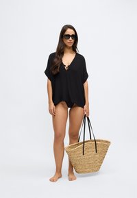 Črni kaftan z izrezom v obliki črke V in rezom, združen z vlisano torbo iz slame z črnimi trakovi. Model stoji bosa in gleda naravnost.