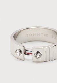 Ασημί δαχτυλίδι Tommy Hilfiger με δύο διάφανα πέτρες, με χαραγμένο το όνομα της μάρκας στο εσωτερικό και διακοσμητικές ρίγες σε κόκκινο, άσπρο και μπλε στην ταινία.