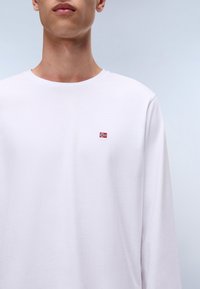 Hvid langærmet t-shirt med rund halsudskæring, der har et lille emblem af det norske flag i rød og blå på brystet. Blødt bomuldsstof.
