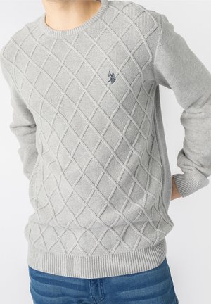Hellgrauer gestrickter Pullover mit Diamantmuster und kleinem dunkel besticktem Logo auf der Brust, getragen zu blauen Jeans.