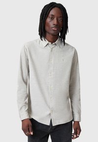 Lys grå langærmet skjorte med knapkrave, struktureret stof og subtil logo-brodering på brystet, kombineret med mørke jeans.