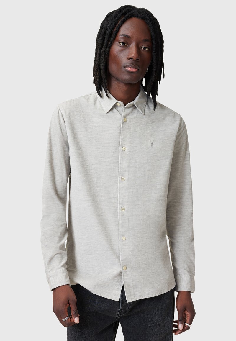 Lys grå langærmet skjorte med knapkrave, struktureret stof og subtil logo-brodering på brystet, kombineret med mørke jeans.