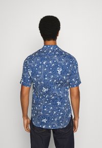 En man visas bakifrån iklädd en kortärmad blå skjorta med ett vitt mönster, tillsammans med mörka jeans, mot en enfärgad bakgrund.