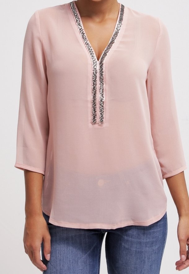 Blouse rose clair à manches 3/4, col en V, bordure de sequins argentés, portée avec un jean bleu sur une personne debout devant un fond blanc.