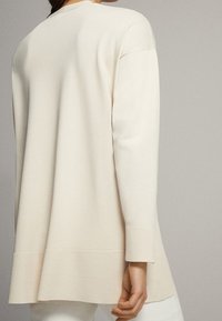 Frau trägt einen locker sitzenden, langärmligen, hellbeigen Pullover mit gerippten Bündchen und Saum, von hinten vor einem einfarbigen Hintergrund gezeigt.