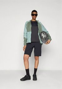 Lightweight cycling jacket in teal and grey, paired with a black t-shirt and shorts. Black sneakers and helmet complete the outfit.