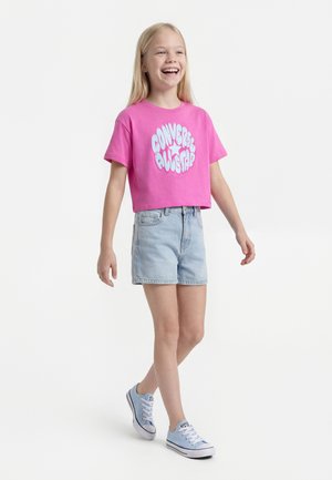 Fille souriante marchant, portant un t-shirt court rose Converse All Star, un short en jean clair et des baskets bleu clair.