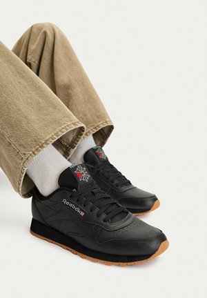 Persona con zapatillas negras de cuero Reebok con suelas de goma, calcetines blancos y pantalones beige contra un fondo claro y liso.