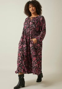 Robe longue noire avec un motif floral rose et rouge, taille cintrée, manches longues et poches latérales. Associée à des bottes noires jusqu'aux genoux.