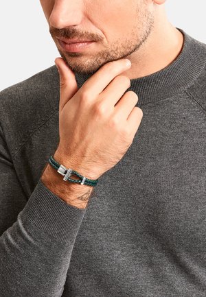 Mann mit kurzem Bart, der einen dunkelgrauen Pullover trägt, berührt sein Kinn und trägt ein grünes geflochtenes Lederarmband mit silberner Schließe am Handgelenk.