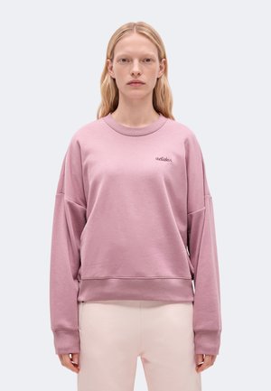 Модел стои на изчистен фон, облечен в розов суичър Adidas и светли панталони.