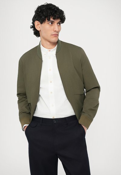 Oliiviroheline pommitaja jope soonikkoes mansettidega, tõmblukuga, ees taskud, kantud valge nööpidega särgi ja tumedate pükste peal.