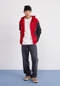 Rød og sort letvægtsjakke med hætte, med en hvid accentlinje og logo. Hvid t-shirt, mørke jeans og multikolorede sneakers.
