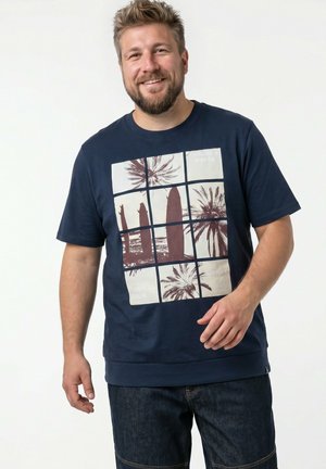 Uśmiechnięty mężczyzna z krótkimi jasnobrązowymi włosami i brodą, ubrany w granatowy t-shirt z brązowym wzorem kratki przedstawiającym palmy i deski surfingowe oraz ciemne dżinsy.