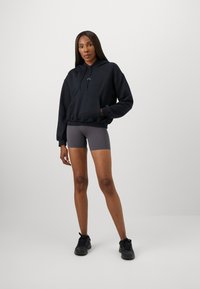 Hoodie noire avec une poche avant, associé à un short de cyclisme gris. Le mannequin porte des sneakers noires, mettant en valeur un look décontracté et athlétique.