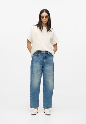 Kvinde iført beige oversized t-shirt, blå højtaljede vide jeans, hvide loafers og små solbriller med gultonet glas, stående med hænderne bag ryggen.