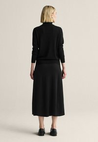 Nainen lyhyillä vaaleilla hiuksilla, pukeutunut mustaan pitkähihainen paitaan, mustaan midihameeseen ja mustiin kenkiin, seisoo selin yksinkertaisella taustalla.