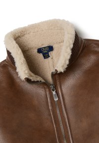 Chaqueta de cuero marrón con un cuello de borreguito sintético crema, cierre de cremallera y una etiqueta que dice "OVS kids, años 4-5." Textura suave, diseño clásico.