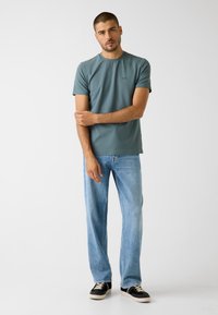 T-Shirt in Türkis mit kurzen Ärmeln und Rundhalsausschnitt, kombiniert mit hellblauen Jeans und schwarzen Turnschuhen. Glatter Stoff, minimalistisches Design.