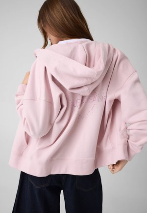 Jeune femme de dos portant un sweat à capuche rose clair avec texte en strass dans le dos et un jean foncé à jambes larges.