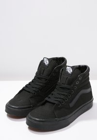 Încălțăminte sport neagră în stil high-top, confecționată din pânză, cu o suprafață texturată, cu linia iconică Vans, șireturi rotunde și ochiuri metalice.