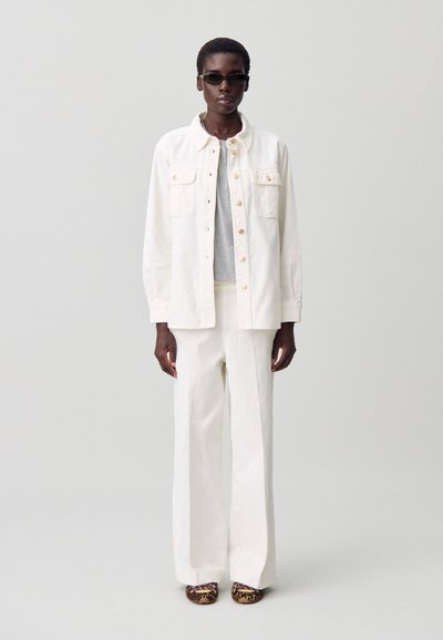Giacca di jeans bianca con bottoni dorati e due tasche sul petto, abbinata a pantaloni larghi dello stesso colore e una maglietta grigia; scarpe con stampa animale completano il look.