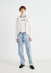 Cardigan blanco de punto acanalado con capucha, llevado sobre una camiseta blanca ajustada con logotipo negro, combinado con jeans holgados de color azul claro y zapatos negros.