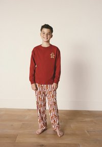 Cămașă roșie cu mânecă lungă, cu broderie de om de turtă dulce, combinată cu pantaloni de pijama albi cu modele cu dungi roșii și desene de fursecuri de turtă dulce.
