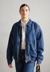 Veste en denim avec fermeture éclair, coupe oversize, couleur bleu foncé et poignets élastiques. Présente un patch logo à l'avant.