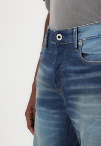 Blå denimjeans med ett blekt mönster, med en klassisk femficksdesign, knäppning och kontrasterande sömnadsdetaljer.