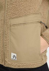 Beige fleecejakke med en struktureret overflade og glatte stofflader. Har en lynlås lomme og et lille logo mærke på fronten.