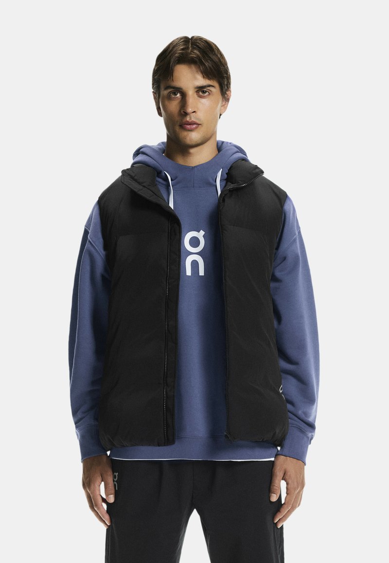 Chaleco negro acolchado con capucha, cremallera frontal y bolsillos laterales, llevado sobre un sudadera azul con logo blanco. Textura suave, ajuste casual.