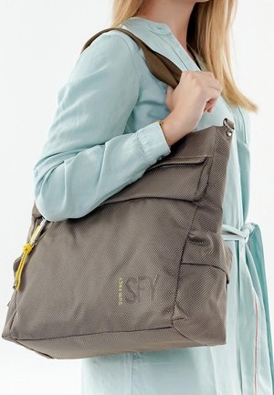 Donna che indossa una camicia azzurro chiaro e tiene una grande borsa a tracolla marrone con texture, con una cerniera gialla e molteplici tasche.