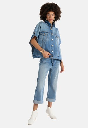 Giacca di denim azzurro oversize con bottoni a pressione, due tasche frontali e colletto alto, abbinata a jeans azzurri a sigaretta e stivaletti bianchi alla caviglia.