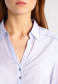 Chemise bleu clair à boutons avec un motif géométrique subtil, tissu doux et col classique. Comprend deux boutons gris à l'avant.