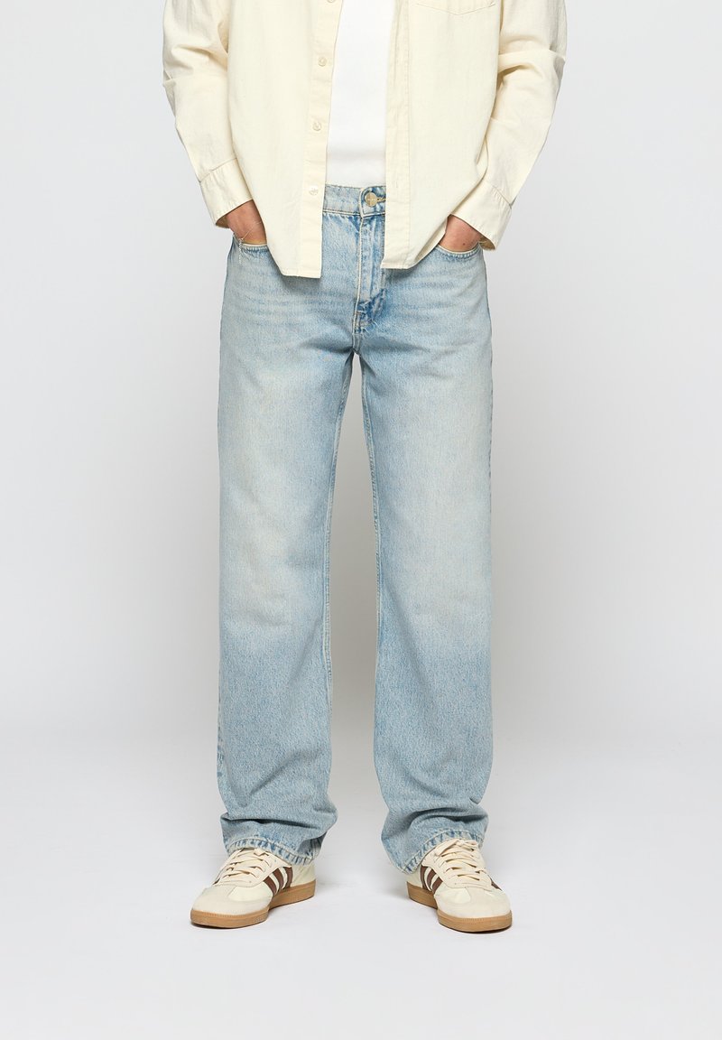 Light blue denim jeans with a relaxed fit, featuring traditional five-pocket styling and subtle fading. Worn with a cream button-up shirt and sneakers.