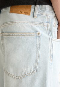Shorts en denim bleu clair avec une pièce en cuir marron portant l'étiquette "PULL&BEAR", présentant des coutures visibles et une texture usée près de la poche.