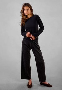 Donna in un top nero a maniche lunghe e pantaloni larghi scintillanti posa in piedi contro uno sfondo grigio uniforme.
