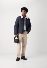 Mand iført en navy pufferjakke med hvid fleecekrave, beige bukser, sorte sko, der holder en lille sort taske, stående mod en ensfarvet hvid baggrund.