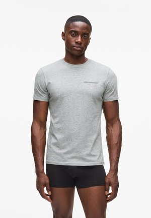 Rövid hajú férfi világosszürke testhezálló pólóban és fekete rövidnadrágban, sima fehér háttér előtt áll.