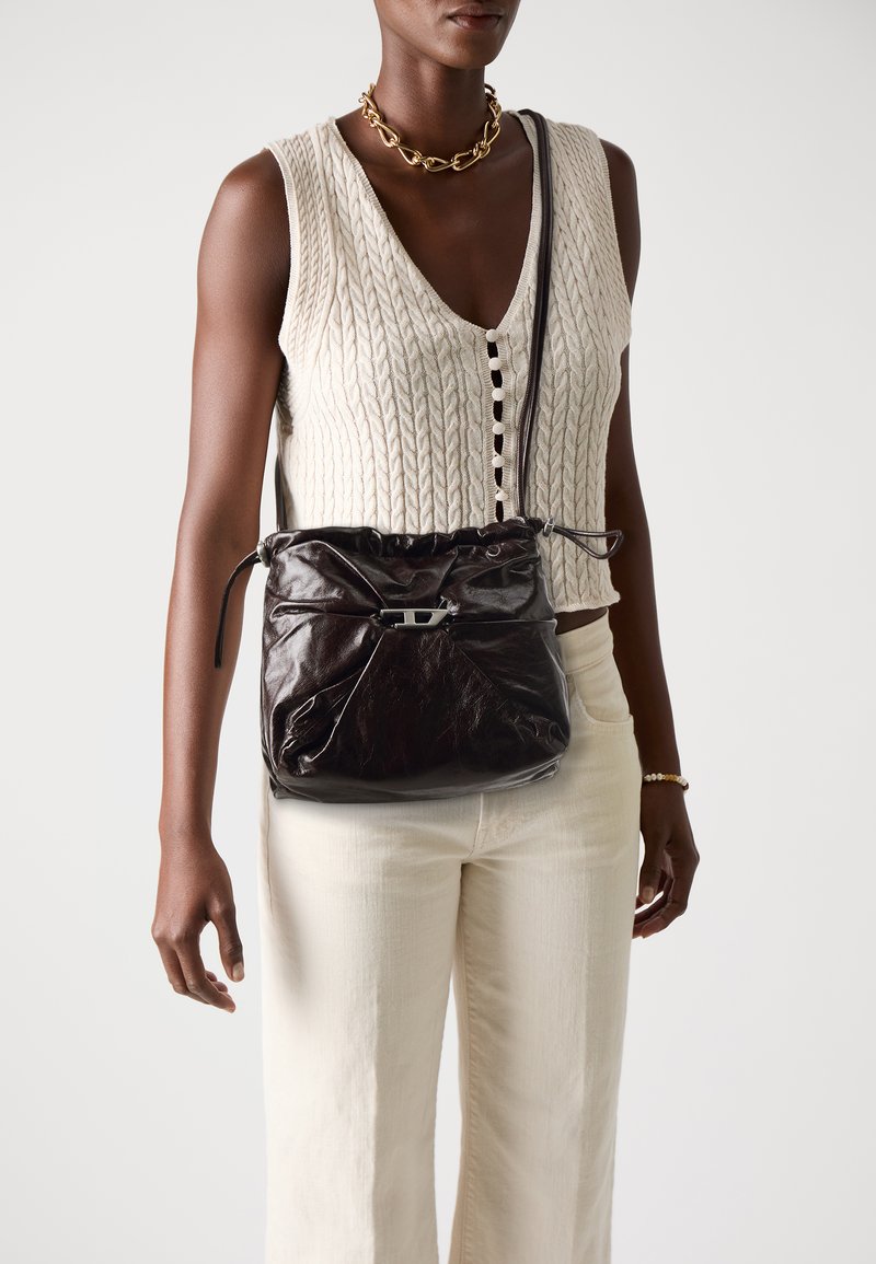Woman wearing cream cable-knit sleeveless vest and pants with chunky gold necklace and dark brown leather crossbody bag.