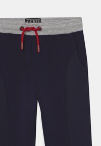 Marina blåa sweatsbyxor med ett grått elastiskt midjeband, röd dragsko, texturerade sidopaneler och GUESS-logotyp på midjebandet.