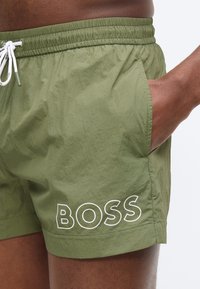 Πλάγια όψη ατόμου που φοράει μαγιό σε ελιά πράσινο με λευκό κορδόνι και λογότυπο "BOSS" στο αριστερό πόδι, με το χέρι στην τσέπη.