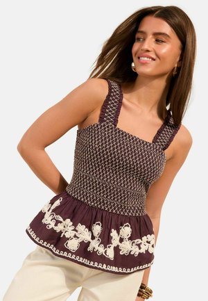 Femme portant un haut sans manches violet foncé à motifs avec une broderie florale blanche et un ourlet volanté, associé à un pantalon clair.