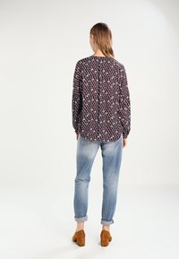 Camicia a maniche lunghe in tessuto scuro con un motivo geometrico multicolore, abbinata a jeans in denim chiaro e scarpe con tacco marroni.