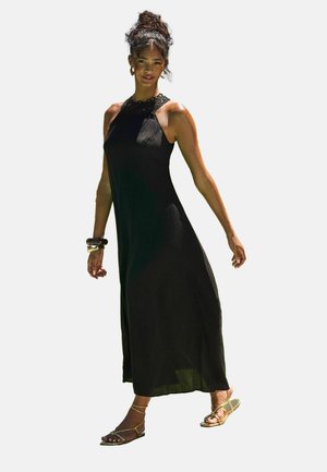 Robe maxi noire avec un col halter et un tissu texturé, ornée de perles délicates au niveau du décolleté. Portée avec des sandales plates à accents dorés.