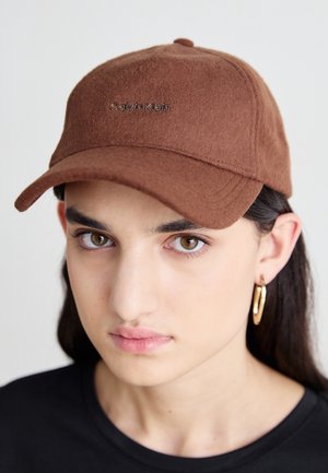 Młoda osoba o brązowych oczach, nosząca brązową czapkę Calvin Klein oraz złote kolczyki koła na jednolitym tle.