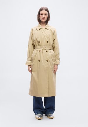 Kvinde iført beige dobbeltradet trenchcoat med bælte, vide jeans og sølvfarvede sneakers stående foran en ensfarvet hvid baggrund.