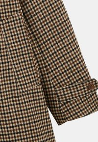 Tissu en pied-de-poule brun et noir avec une texture lisse ; présente un détail de poignet boutonné et un design de col pointu.